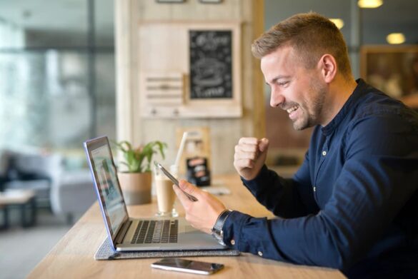 طراحی مجدد وب سایت، یک فرد خوشحال که تلفن همراه در دست دارد پشت یک میز در یک کافه که بر روی آن یک لپ تاپ قرار دارد نشسته است